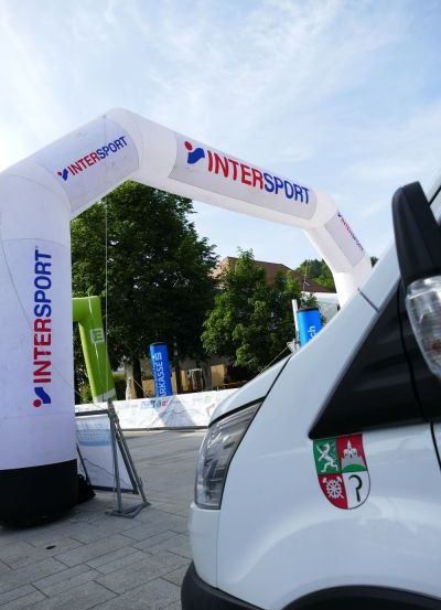 Ein weißer Van ist unter einem aufblasbaren Bogen mit dem INTERSPORT-Logo geparkt, mit Bäumen und einem Gebäude im Hintergrund.
