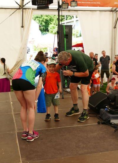 Ein Mann und eine Frau auf der Bühne mit einem jungen Jungen dazwischen. Sie erhalten eine Medaille. Im Hintergrund ist eine Menge.