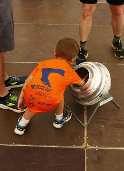Ein junger Junge versucht, einen großen Metallfass zu drehen, wobei zwei Erwachsene hinter ihm stehen. Der Junge trägt ein orangefarbenes Shirt mit einem blau-weißen Logo.