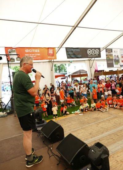 Ein Mann steht auf einer Bühne und spricht in ein Mikrofon vor einem Publikum. Die Bühne ist mit Lautsprechern und Bannern ausgestattet. Das Publikum besteht aus Erwachsenen und Kindern, von denen viele orangefarbene Shirts tragen.