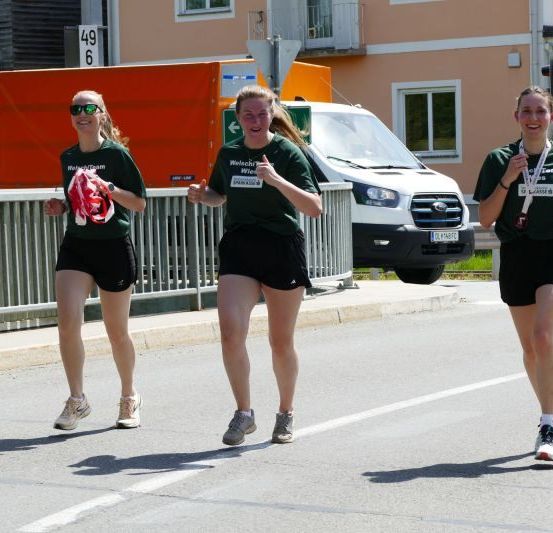 Drei Frauen in passenden grünen T-Shirts und schwarzen Shorts laufen in einem Rennen und halten rote Bänder. Sie sind auf einer Straße mit einem Metallzaun und Gebäuden. Ein weißer Van ist in der Nähe geparkt.