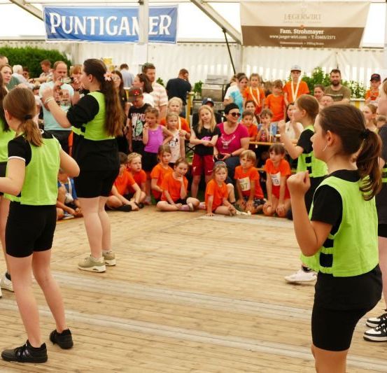 Eine Gruppe junger Menschen in grünen Westen tritt auf einer Bühne auf, während viele Menschen zusehen. Einige tragen orangefarbene Shirts, und es gibt Banner mit Text hinter den Darstellern.