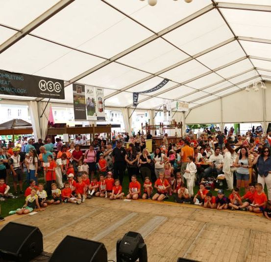Eine große Menschenmenge, darunter auch Kinder, versammelt sich unter einem weißen Zelt. Viele sitzen auf dem Rasen, während andere stehen, einige tragen orangefarbene Shirts. Es gibt eine Bühne mit Lautsprechern und Bannern, die am Zelt hängen.