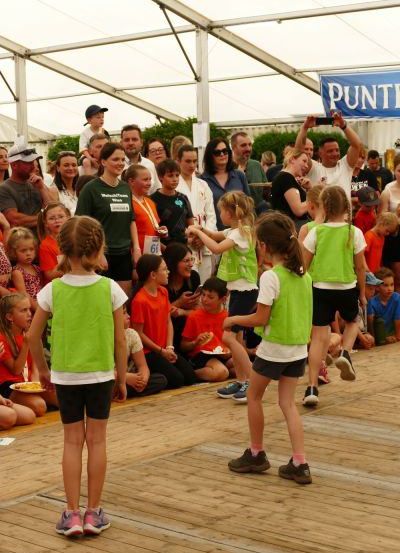 Eine Gruppe von Kindern in grünen Westen tritt auf einer Bühne unter einem Zelt auf, während eine Menge zuschaut. Ein Banner mit dem Wort 'PUNT' ist sichtbar.