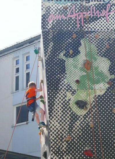 Ein Kind klettert mit einem Sicherheitsgurt an einer Wand. Die Wand ist mit verschiedenen Farben und Designs verziert. Das Kind hält sich mit beiden Händen an der Wand fest.