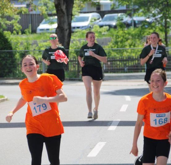 Eine Gruppe junger Frauen in orangefarbenen Shirts läuft in einem Marathon auf einer Stadtstraße. Bäume und geparkte Autos säumen die Seiten.