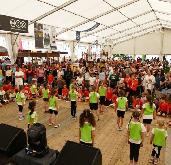 Eine große Menschenmenge versammelt sich unter einem weißen Zelt für eine Veranstaltung. Kinder in grünen und orangefarbenen Westen treten auf einem Holzboden auf, während Erwachsene um sie herum stehen und sitzen. Plakate und Lautsprecher sind vorhanden.