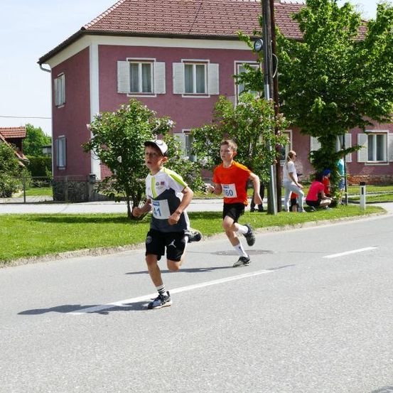 Zwei Jungen rennen auf einer Straße mit den Nummern 60 und 11. Ein Haus und ein Baum sind hinter ihnen. Einige Leute stehen in der Nähe des Hauses.