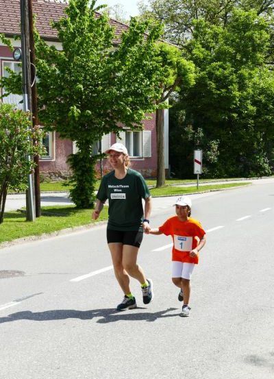 Eine Frau und ein Kind laufen ein Rennen und tragen Nummern auf ihren Trikots. Sie sind auf einer Straße mit Häusern und Bäumen.