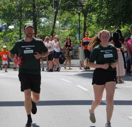Zwei Läufer nehmen an einem Marathon teil, während andere hinter ihnen laufen. Die Läufer tragen grüne Shirts mit weißen Schriftzügen. Zuschauer säumen die Straße und beobachten die Läufer.