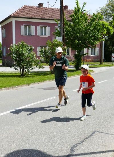 Ein Mann und ein junges Mädchen laufen auf einer Straße mit einem roten Haus und Bäumen im Hintergrund.
