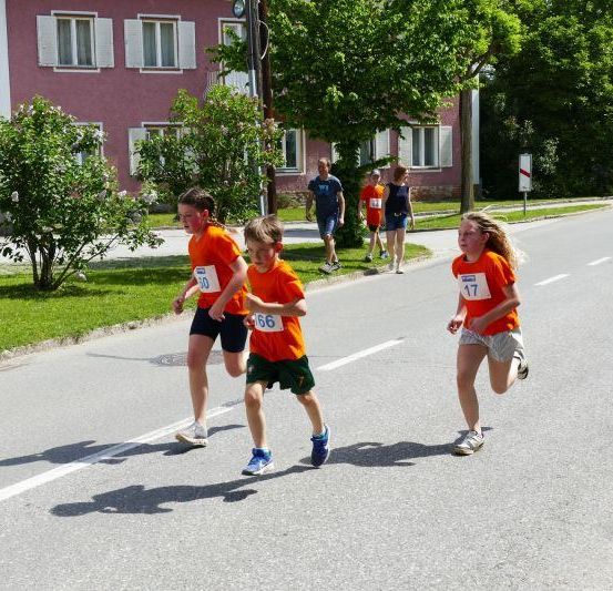 Drei Kinder laufen auf der Straße. Sie tragen die Nummern 60, 66 und 17. Dahinter laufen zwei Personen auf dem Bürgersteig. Auf der linken Seite befindet sich ein Haus mit weißen Fensterläden und einem Baum. Auf der rechten Seite steht ein weiterer Baum und ein Pfosten.
