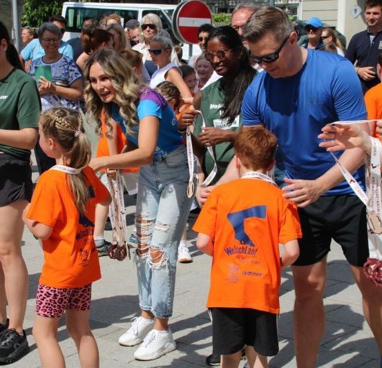 Mehrere Erwachsene und Kinder in orangefarbenen T-Shirts versammeln sich um eine Gruppe von Menschen in grünen und blauen Shirts. Sie tragen alle Sonnenbrillen und einige halten Medaillen. Die Menschen in Blau und Grün verteilen Medaillen an die Menschen in Orange. Im Hintergrund steht eine Menschenmenge.