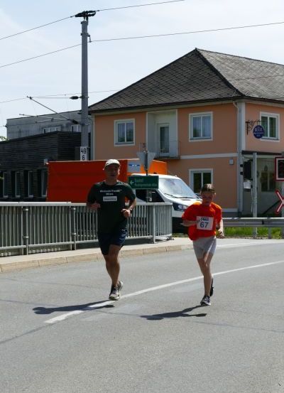 Zwei Männer laufen auf einer Straße, einer trägt ein schwarzes Shirt mit einer Nummer und der andere ein orangefarbenes Shirt mit der Nummer 67. Eine Metallbarriere befindet sich an der Seite, und ein Gebäude mit Fenstern steht dahinter.