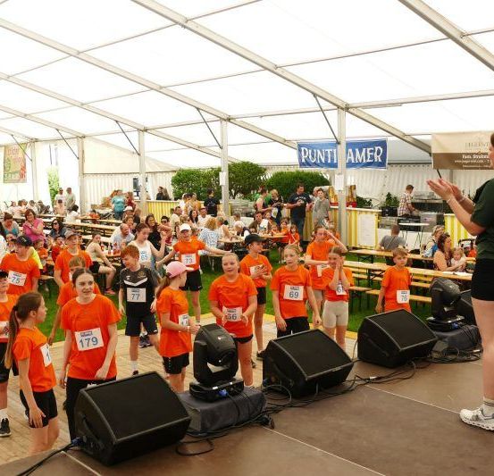 Eine Gruppe von Kindern in orangefarbenen Shirts steht auf einer Bühne unter einem Zelt. Eine Frau spricht zu ihnen. Das Publikum sitzt im Hintergrund.