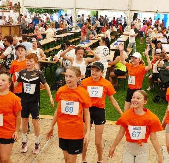 Eine Gruppe junger Menschen in orangefarbenen T-Shirts mit Nummern tanzt in einem Außenbereich mit Tischen und sitzenden Personen.