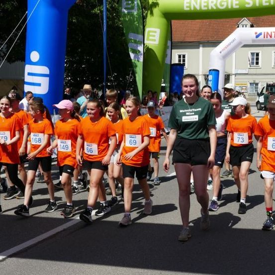 Eine Gruppe von Kindern und Erwachsenen nimmt an einem Laufevent teil, mit nummerierten orangefarbenen T-Shirts. Eine Frau in Grün führt sie an. Im Hintergrund befindet sich ein dekorativer Bogen und Gebäude.