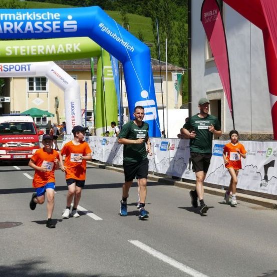 Teilnehmer eines Marathons, die durch eine Stadt laufen, mit einem großen Bogen, auf dem 'Steiermarkische Sparkasse' und 'Intersport' steht. Zuschauer und ein roter Krankenwagen im Hintergrund.