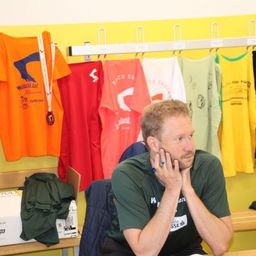 Ein Mann sitzt in einem Raum mit farbenfrohen T-Shirts und Medaillen, die hinter ihm hängen. Er trägt ein grünes Shirt mit dem Wort 'HSS' darauf.