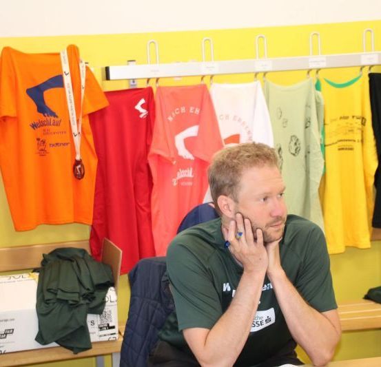 Ein Mann sitzt in einem Raum mit farbenfrohen T-Shirts und Medaillen, die hinter ihm hängen. Er trägt ein grünes Shirt mit dem Wort 'HSS' darauf.