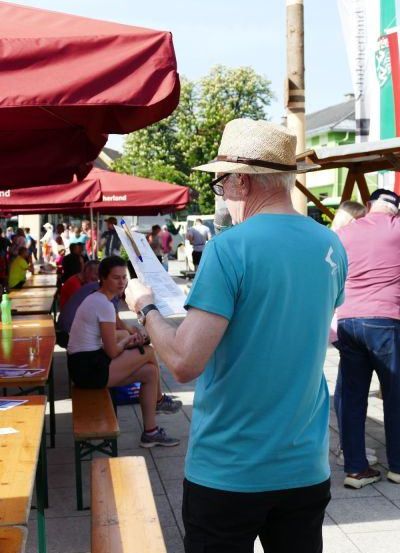 Ein älterer Mann in einem Hut steht auf einem Freimarkt, hält ein Stück Papier und einen Stift in der Hand, umgeben von Menschen, die an Tischen sitzen.