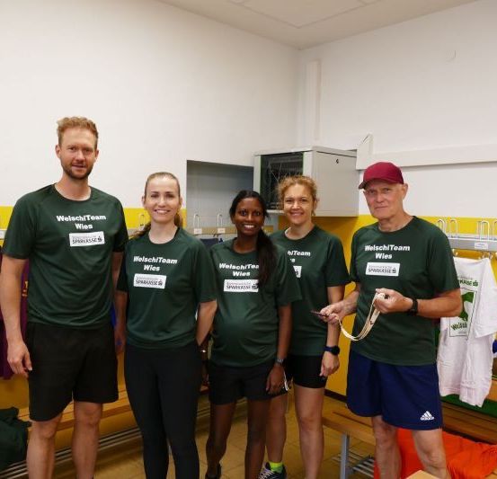 Fünf Personen stehen in einem Raum und tragen grüne T-Shirts mit dem Text 'WelschTeam Wies' darauf. Sie lächeln alle und posieren für ein Foto. Hinter ihnen befinden sich Kleiderständer und eine gelbe Wand.