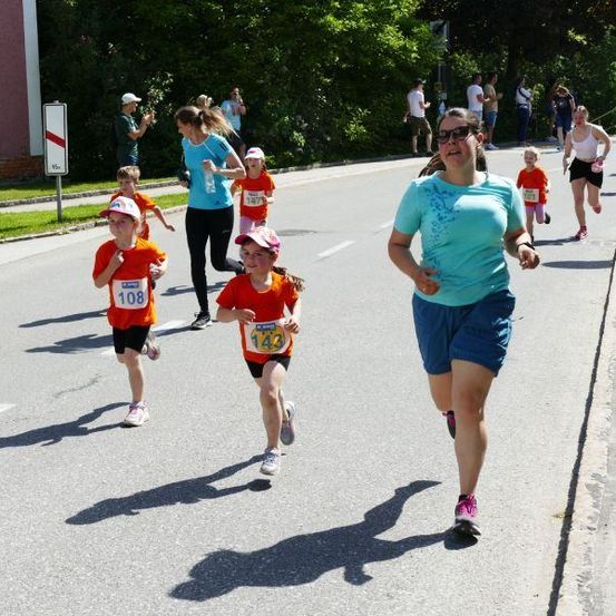 Eine Gruppe von Menschen läuft bei einem Marathon. Zu den Läufern gehören eine Frau, ein junges Mädchen und Kinder mit Nummern auf ihren Trikots. Einige Leute stehen am Straßenrand.