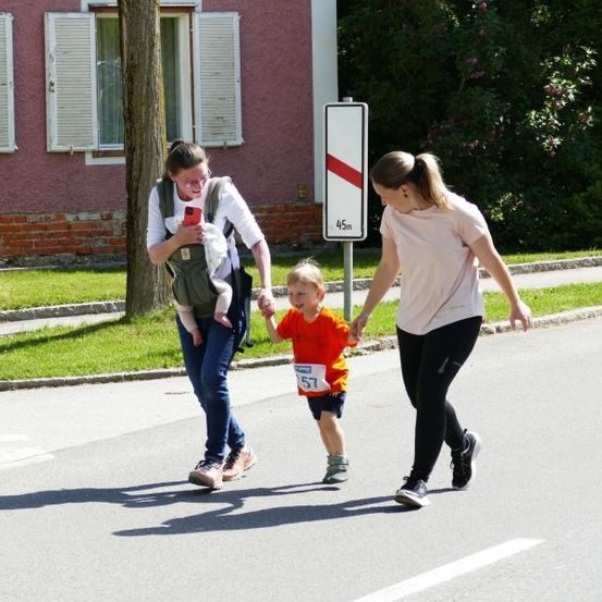 Eine Familie geht mit einem Baby in einem Tragegurt und einem kleinen Jungen, der die Hand seiner Mutter hält, über die Straße.