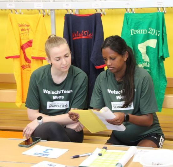 Zwei Frauen sitzen an einem Schreibtisch und überprüfen Papiere. Eine hält eine Mappe, die andere einen Marker. Sie tragen grüne T-Shirts mit der Aufschrift 'WelschTeam'. Hinter ihnen hängen Kleidungsstücke an einem Ständer mit T-Shirts, auf denen 'Team 2017' steht.