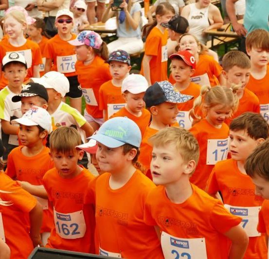 Eine Gruppe von Kindern, die orangefarbene Trikots mit Nummern tragen, steht in einer Reihe, wahrscheinlich im Wartemodus auf den Start eines Rennens.
