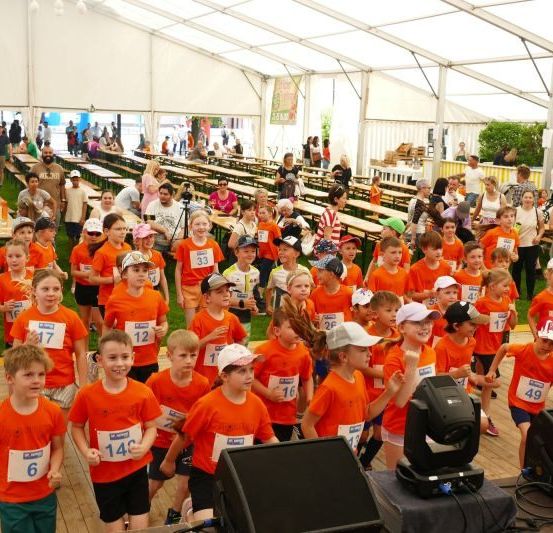 Eine Gruppe von Kindern in orangefarbenen T-Shirts mit Nummern darauf läuft ein Rennen. Die Kinder tragen Mützen und Turnschuhe. Dahinter stehen Holzbänke und Tische und ein weißes Zelt.