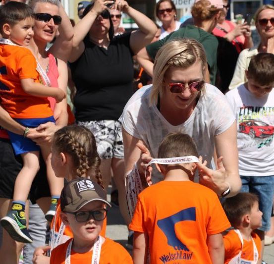 Eine Gruppe von Menschen versammelt sich im Freien. Eine Frau legt einem Jungen ein weißes Band um den Hals. Mehrere Kinder und Erwachsene tragen orangefarbene T-Shirts mit weißen Buchstaben. Einige Leute halten Kameras. Der Hintergrund ist unscharf.