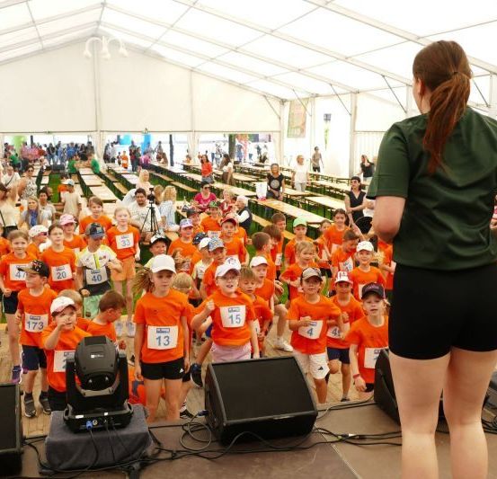Eine Gruppe von Kindern in orangefarbenen Trikots mit Nummern nimmt an einem Rennen in einem großen Zelt teil. Eine Frau in Grün ist auf der Bühne.