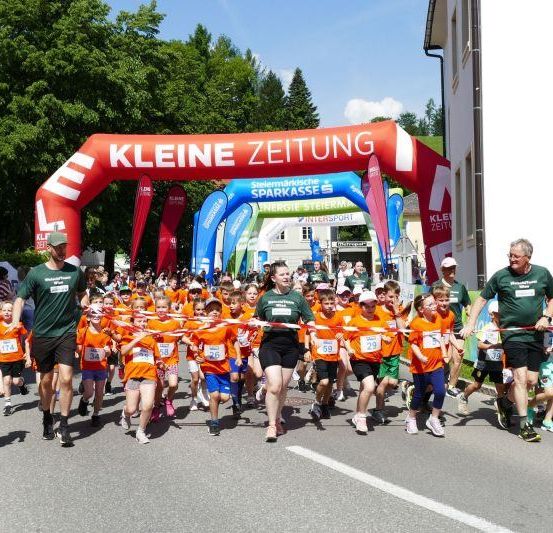 Kinder in orangefarbenen Trikots nehmen an einem Rennen teil und passieren einen roten Bogen mit der Aufschrift 'Kleine Zeitung'.