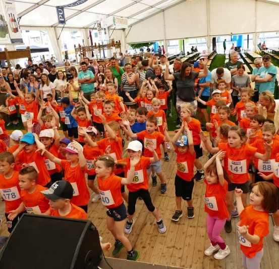 Eine große Gruppe von Kindern in orangefarbenen Shirts mit Nummern jubelt bei einer Veranstaltung, während Erwachsene um sie herum sind. Die Kinder tanzen auf der Bühne.