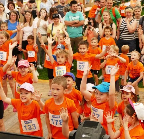Eine Gruppe von Kindern, die orangefarbene Hemden und nummerierte Etiketten tragen, winken fröhlich in einer Außenumgebung. Zuschauer im Hintergrund halten den Moment fest.