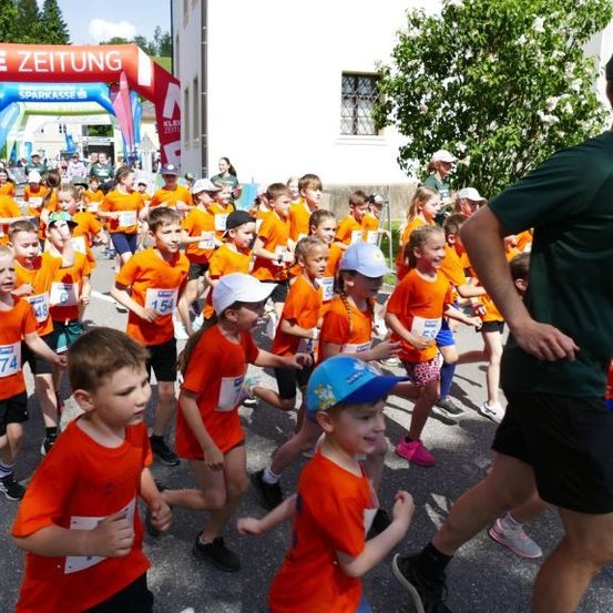 Kinder in orangefarbenen Shirts laufen in einem Marathon mit einem Mann in Grün, der ihnen folgt. Der Bogen hat den Text 'Kleine Zeitung' und 'Sparkasse'.
