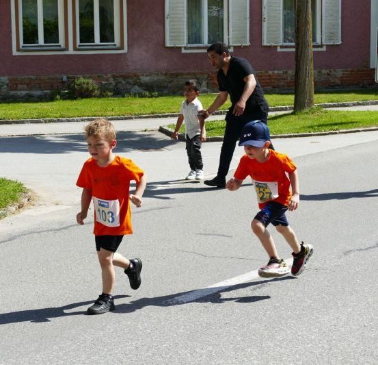 Zwei junge Jungen laufen in einem Rennen, einer mit der Nummer 103 auf seinem Shirt. Ein Mann und ein weiterer Junge beobachten. Eine Frau in Weiß läuft auf der Straße.