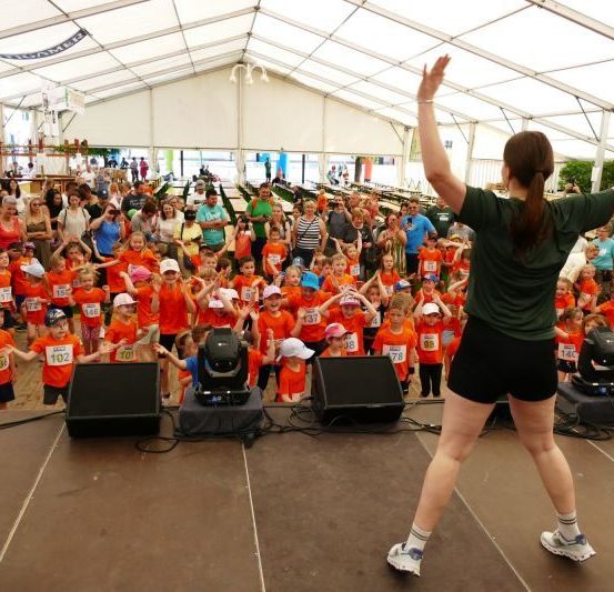 Eine Frau führt eine Gruppe von Kindern in orangefarbenen T-Shirts in einem großen Zelt an. Die Kinder nehmen an einem Rennen teil, mit Nummern auf ihren Trikots. Das Zelt ist mit Zuschauern gefüllt.