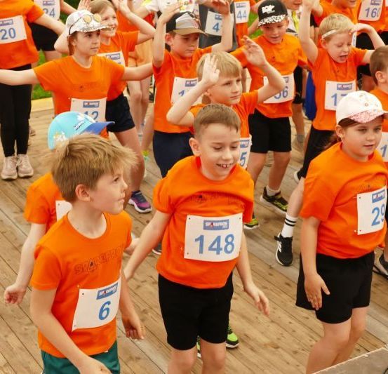 Eine Gruppe junger Kinder in orangefarbenen T-Shirts und Nummern nimmt an einer Veranstaltung auf einem Holzboden teil.