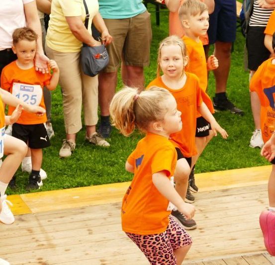 Eine Gruppe von Kindern in orangefarbenen Shirts und Shorts steht auf einer Holzbühne, vermutlich bei einem Wettbewerb. Sie schauen nach rechts, und einige tragen Turnschuhe. Eine Frau in gelber Bluse und khakifarbener Hose steht hinter ihnen.