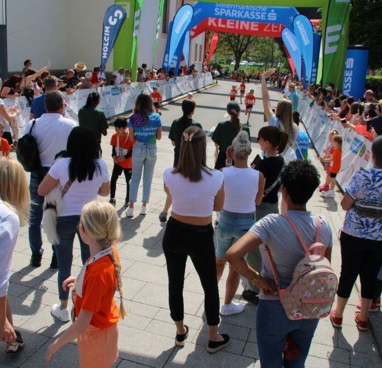 Läufer nehmen an einem Marathon teil, wobei Zuschauer die Strecke säumen. Menschen verschiedenen Alters und Geschlechts sind versammelt, einige tragen Rucksäcke und Kameras. Der Lauf wird von der Sparkasse gesponsert.