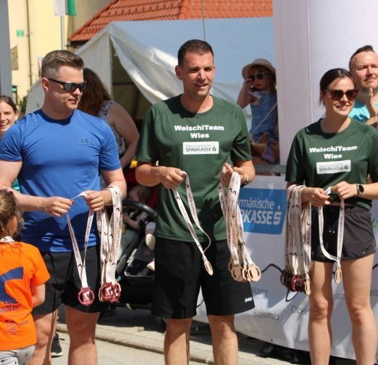 Drei Personen in Sportkleidung stehen zusammen und halten Medaillen. Dahinter versammeln sich Zuschauer, einige machen Fotos. Im Hintergrund ist ein Banner mit rotem und weißem Text zu sehen.