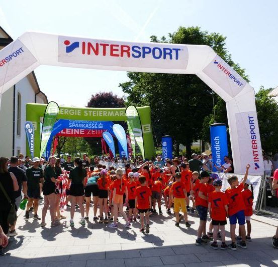 Teilnehmer in orangen Shirts stehen unter einem weißen Intersport-Bogen, mit Zuschauern und Bäumen im Hintergrund.