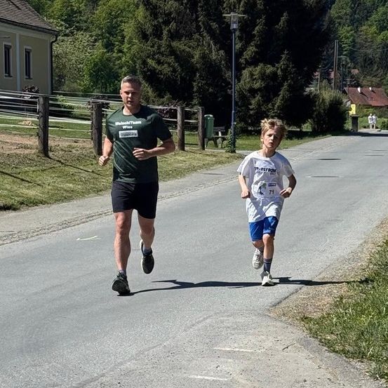 Ein Mann und ein Junge laufen auf einer Straße, in Laufkleidung. Der Junge hat eine Nummer auf seinem Shirt, und sie sind von grünem Gras und Bäumen umgeben.
