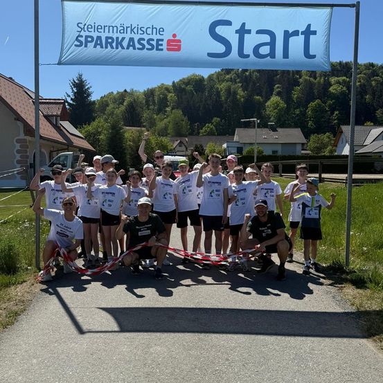 Eine Gruppe von Menschen in weißen T-Shirts posiert an einer Startlinie mit einem Start-Banner darüber. Sie stehen vor Häusern und Grünflächen unter einem klaren blauen Himmel.