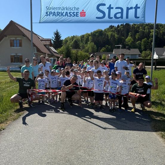 Läufer posieren für ein Foto an der Startlinie eines Rennens. Sie tragen weiße Hemden und Nummern. Ein Banner steht Steiermrkische Sparkasse Start. Dahinter sind Häuser und Grün zu sehen.