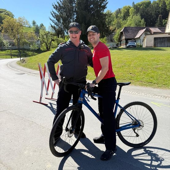 Zwei Männer stehen neben einem blauen Fahrrad auf einer sonnigen Straße, einer in einem schwarzen Jackett, der andere in einem roten T-Shirt. Hinter ihnen sind Häuser, ein Auto und Grünflächen.