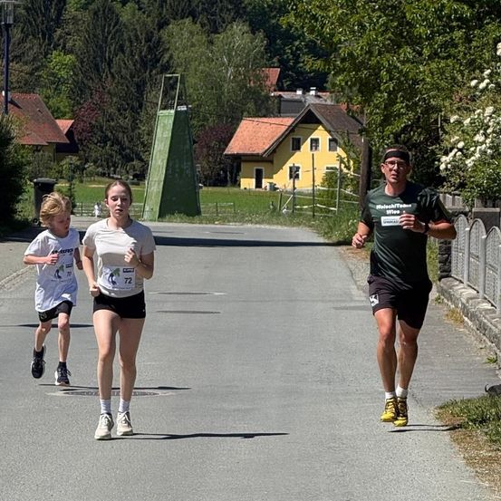 Drei Personen laufen auf einer Straße. Ein Mann ist vorne, gefolgt von einem Mädchen und einem Jungen. Bäume und Häuser sind an der Seite.