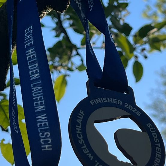 Zwei blaue Bänder hängen von einem Baum, eines mit der Aufschrift Echte Helden Laufen Welsch. Darunter befindet sich eine runde blaue Medaille mit der Aufschrift Finisher 2026 Südsteiermark.